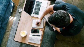 man writing in notebook next to computer